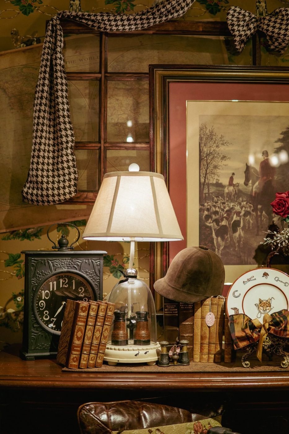 Equestrian vignette — a velvet riding helmet, framed fox-hunt print, leather-bound books and houndstooth ribbon
